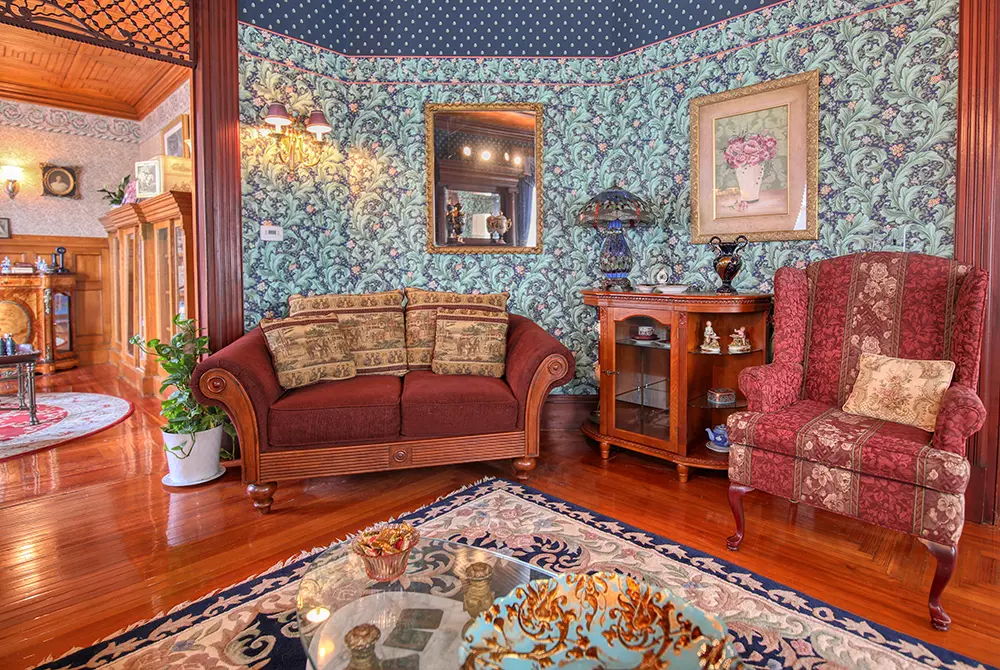 Victorian-style living room with patterned wallpaper, a red sofa and armchair, a wood cabinet, ornate mirror, wall art, and a plant. The polished wood floor and detailed rug complete the elegant, classic decor—perfect for guests seeking a refined stay at a charming bed and breakfast in Toms River NJ.