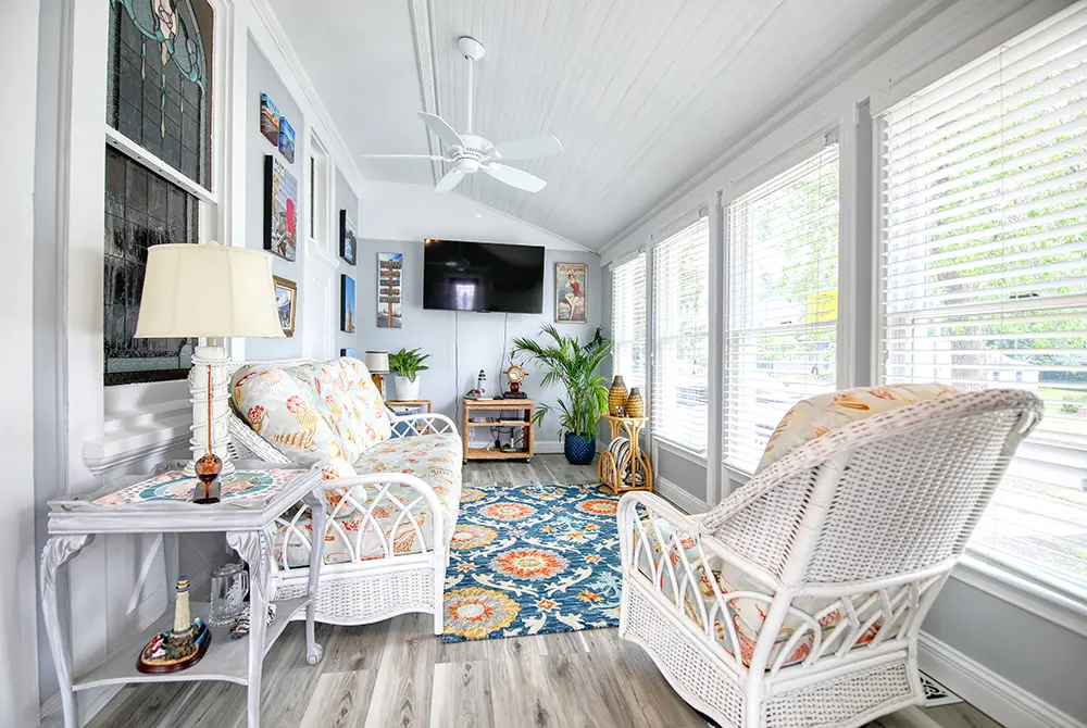 Bright sunroom in a Toms River historic inn, featuring white wicker chairs, a floral rug, plants, and a TV on the far wall. Large windows with blinds let in plenty of natural light, while art, books, and a small lamp add charm to this inviting space.