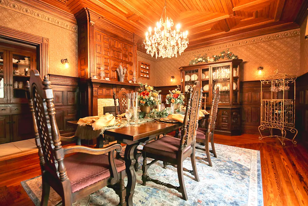 An elegant dining room at this bed and breakfast in Toms River NJ features ornate wood paneling, a chandelier, a large dining table set for a meal, high-backed chairs, a china cabinet, floral centerpiece, and a decorative rug on a polished wooden floor.