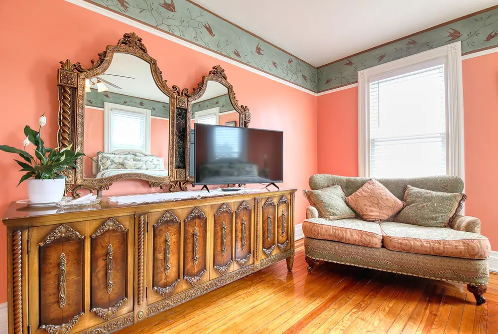 A vintage living room at this charming bed and breakfast in Toms River, NJ features a large ornate wooden cabinet, a TV, two decorative mirrors, a potted plant, a peach-colored loveseat, pink walls, and a bird-patterned border near the ceiling.