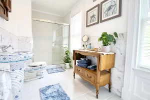 Bright bathroom with a glass shower, blue rugs, plants, and a wooden vanity holding towels and toiletries. Three framed animal sketches hang on the white wall above the vanity—an inviting retreat reminiscent of the charming Toms River NJ bed and breakfast rooms.