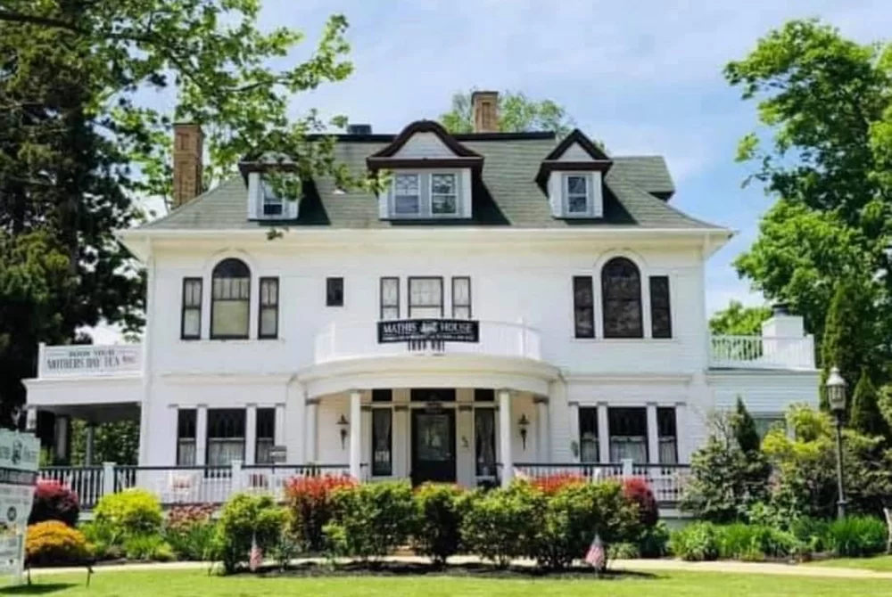 Large white two-story historic house with a mansard roof, dormer windows, and a rounded front porch offers bed and breakfast dining in Toms River, NJ. Surrounded by green trees and bright spring foliage, the property features well-maintained landscaping for a charming and inviting atmosphere.