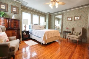 A cozy bedroom with polished wooden floors, a bed with white linens, vintage-style furniture, an armchair, large windows, floral wallpaper, and a ceiling fan with lights.