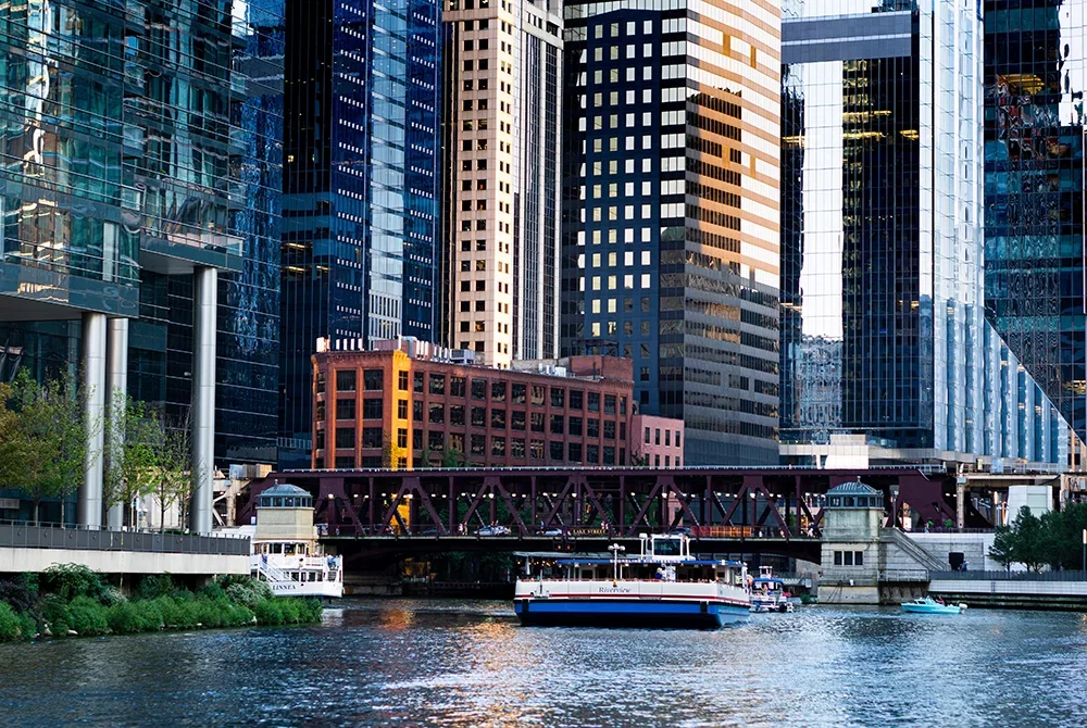 A river boat passes under a bridge surrounded by tall, modern skyscrapers and a red brick building in a cityscape, with greenery along the water’s edge—perfectly capturing the vibrant urban scenery and highlighting some of the best things to do in Toms River NJ.
