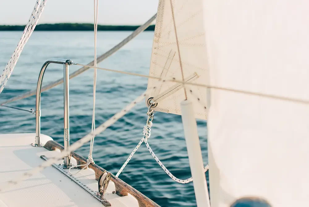 Close-up of a sailboat’s deck and rigging with part of the sail visible, sailing on calm blue water with a distant treeline under a clear sky—a perfect scene for those seeking relaxing things to do in Toms River NJ.
