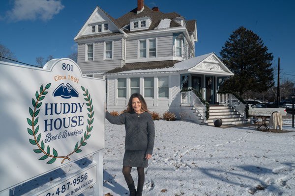 A woman in a gray dress, the Huddy House innkeeper, stands by a Huddy House Bed & Breakfast sign in front of a large white house on a snowy day in Toms River.